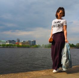 Gina Bley walking on the edge of a lake wearing aubergine coloured pants with a white tshirt that says "Game over"