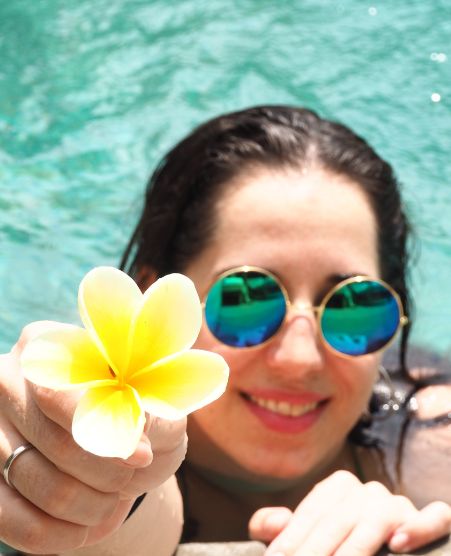 Gina Bley in a pool, wet hair with green-blue sunglasses holding a yellow jepun flower into the camera