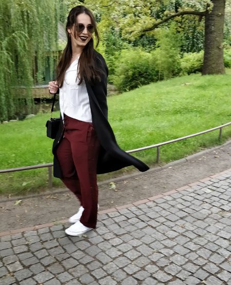 Gina Bley walking in a park, wearing burgundy pants, a long black coat, white shirt and some white shoes