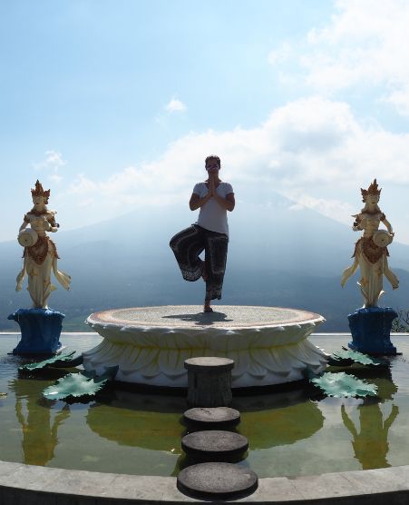 Gina Bley standing in a temple between two statues doing a mountain pose on a platform surrounded by water