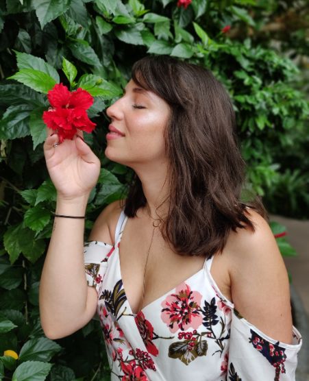 Gina Bley wearing a summery floral dress with cold shoulders, holding a red flower to her face and smelling it with closed eyes