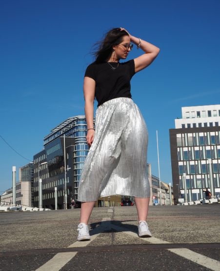 Gina Bley standing in the wind on the street with blue skies in the background, wearing a black tshirt and a silver skirt