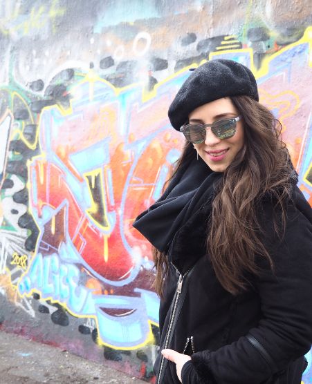 Gina Bley standing in front of graffiti sprayed wall, wearing all black jacket, scarf and beret with silver sunglasses