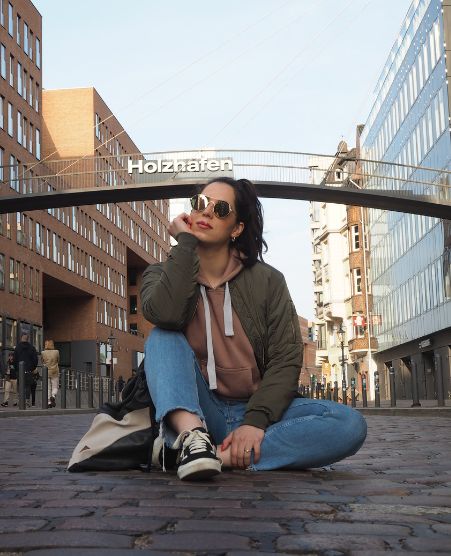 Gina Bley sitting on a cobblestone street, wearing jeans, jumper and bomber jacket with gold sunglasses