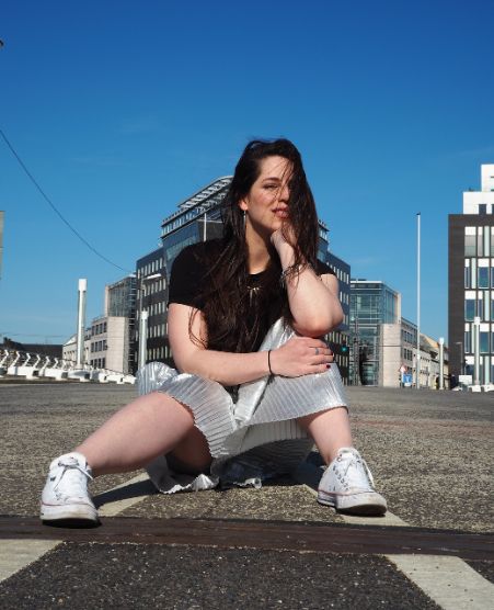 Gina Bley sitting on the street with blue skies in the background, wearing a black tshirt and a silver skirt