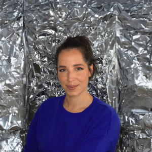 Gina Bley in front of a aluminium backdrop, dressed in a blue jumper with ponytail for the Facebook section