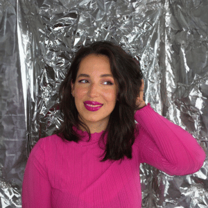 Gina Bley in front of a aluminium backdrop, dressed in a fuchsia pink longsleeved top with pink lipstick for the Instagram section