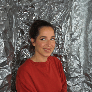 Gina Bley in front of a aluminium backdrop, dressed in a red jumper top with ponytail for the YouTube section