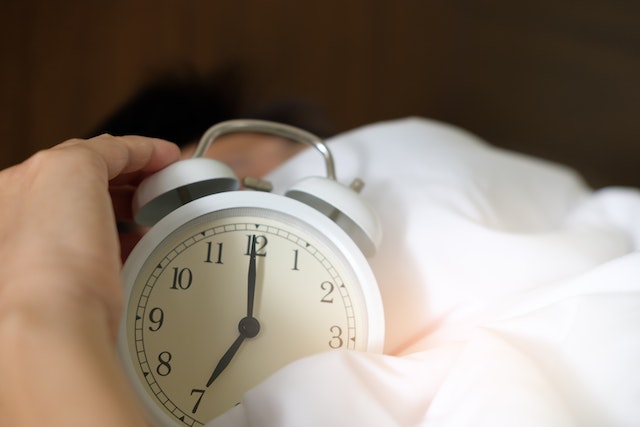 Image for the article "Why Sleep is the Foundation of Change" shows a hand on an alarm clock laying in bed sheets