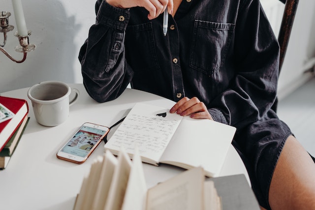 Image for the article "The Power of Journaling for Personal Insight" shows a woman sitting at a table with a journal and her mobile thinking